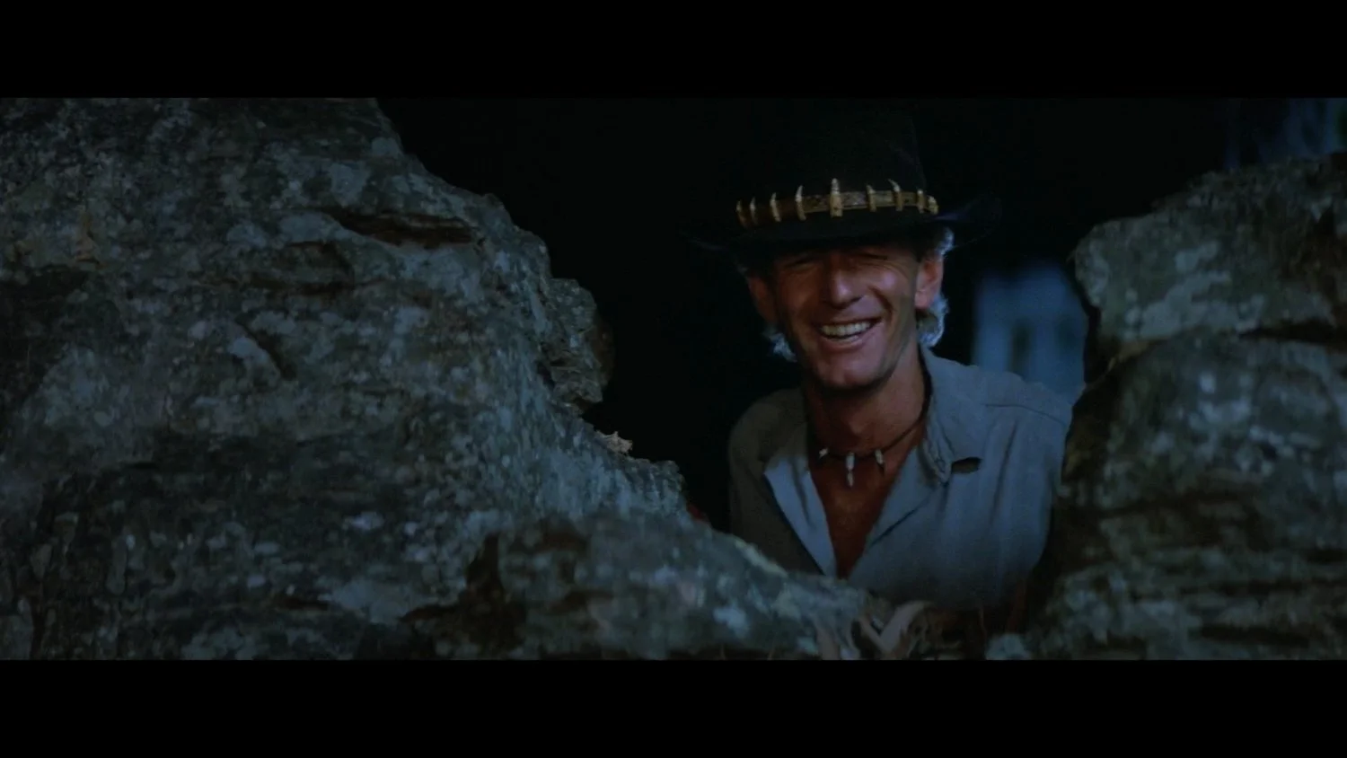 Mick Dundee ( Actor Paul Hogan ), smiling while peeking through a small opening between rocks, wearing his Akubra hat and a necklace with small beads.