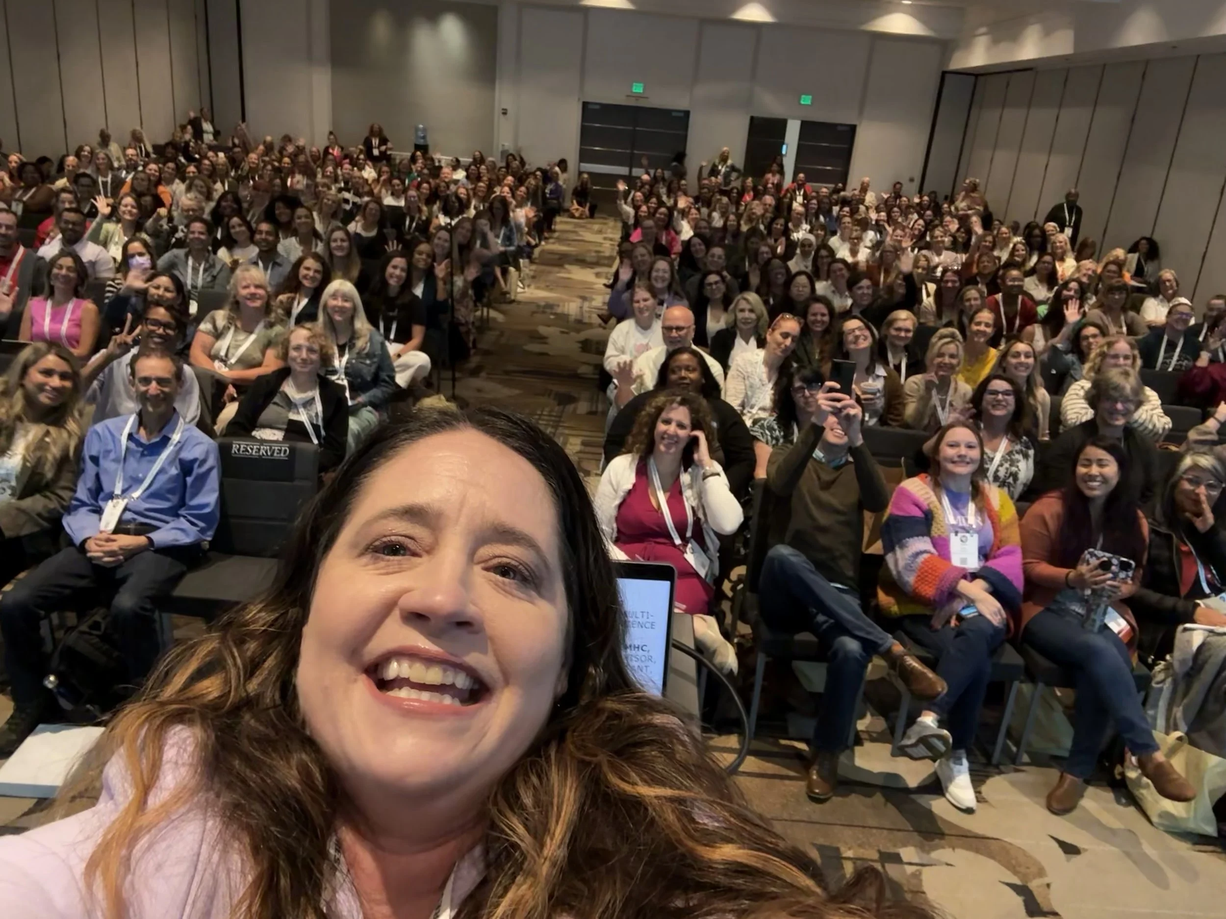 A selfie of a woman with curly hair smiling in front of a large gathering of people seated in rows at a conference or seminar. The audience members are smiling, waving, and looking at the camera with a few holding up peace signs.