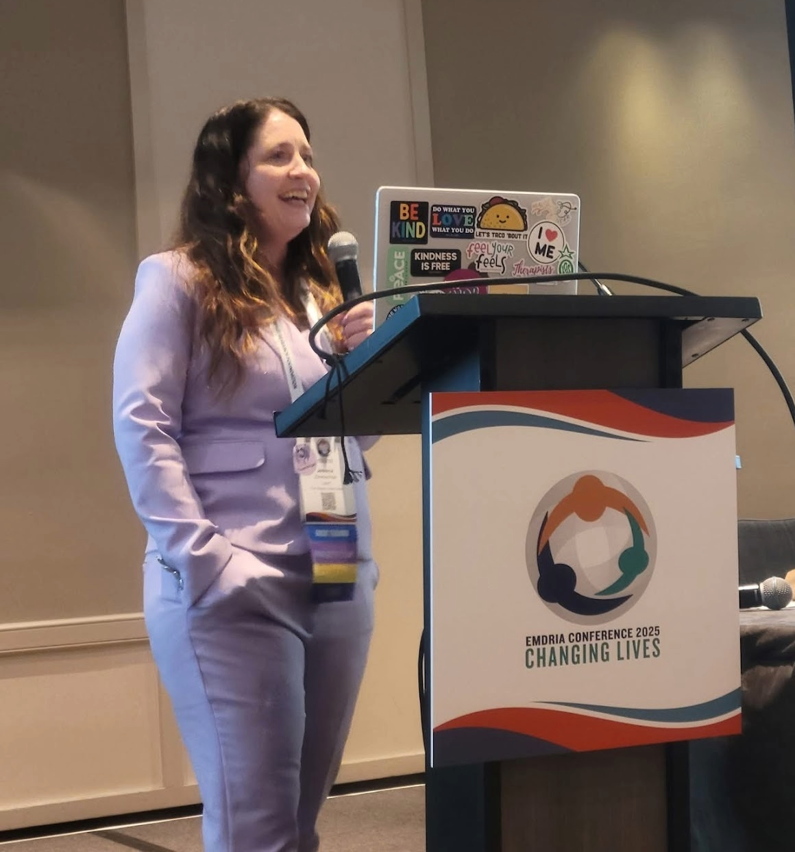A woman in a light pink suit standing at a podium with a microphone, giving a presentation at the EMDRIA Conference 2025 titled 'Changing Lives'. There is a colorful 'Changing Lives' logo on the front of the podium, and her laptop, covered with various stickers, is open on a stand.