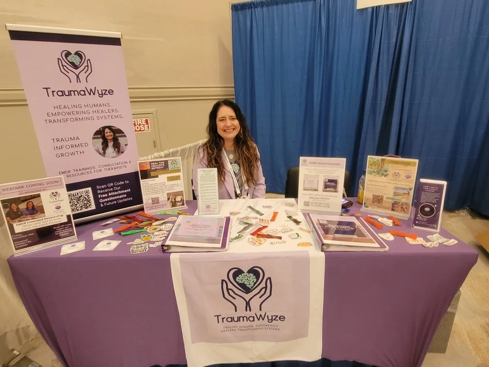A woman sitting at a table with a purple tablecloth and TrauaWye promotional materials, at an indoor event with a blue curtain backdrop.