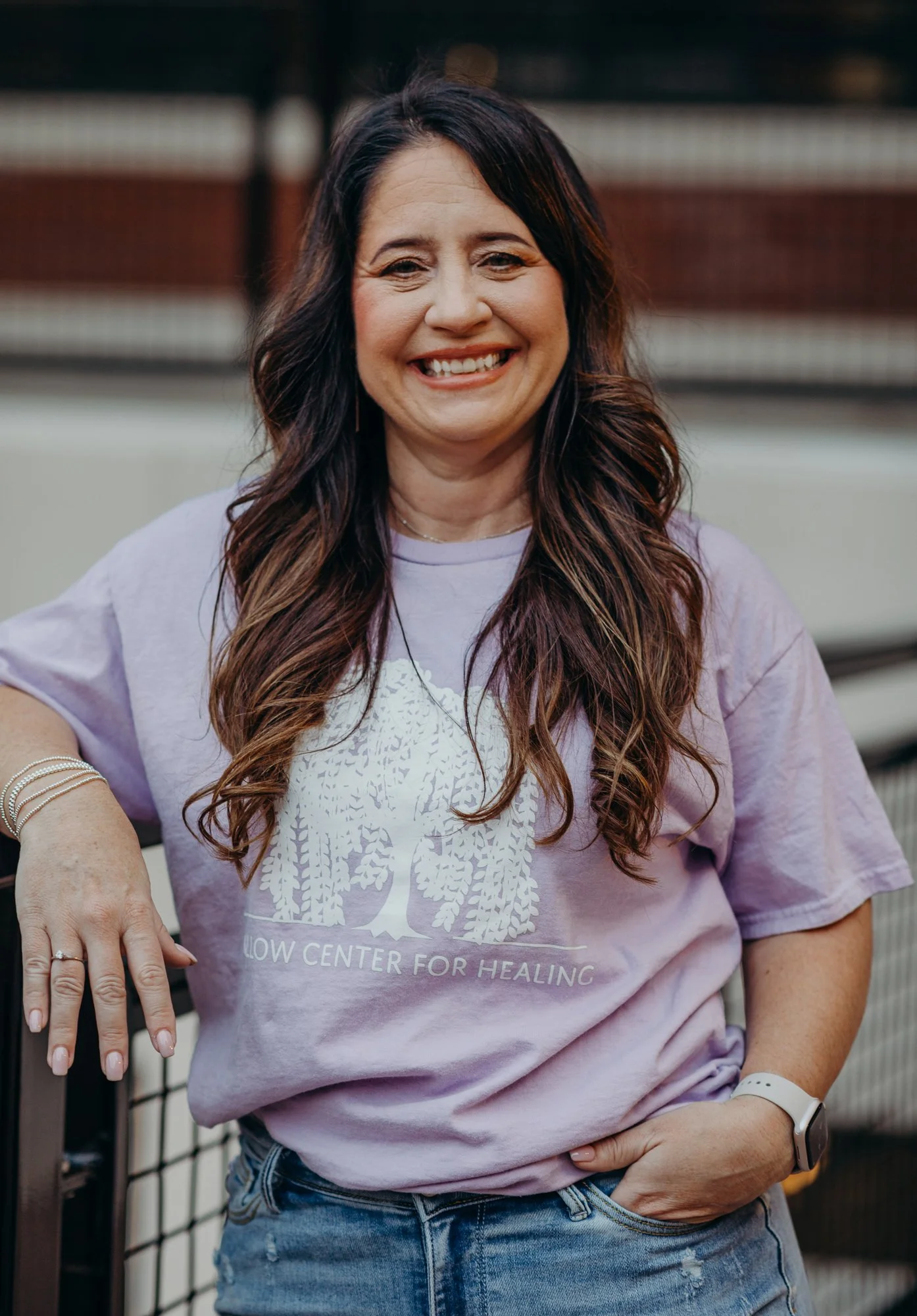 A woman with long brown hair wearing a purple T-shirt with a tree logo and the words 'YELLOW CENTER FOR HEALING' on it. She is smiling and standing outdoors near a fence.
