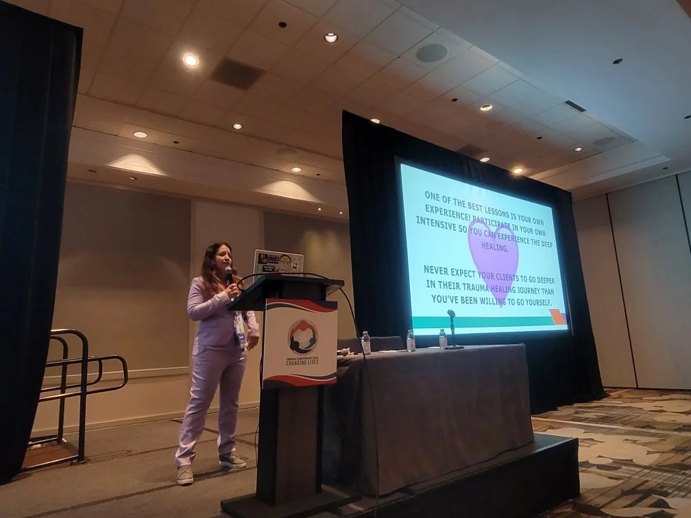 A woman presenting at a conference, standing behind a podium with a screen displaying a slide about lessons on trauma healing, with a table beside her holding water bottles.