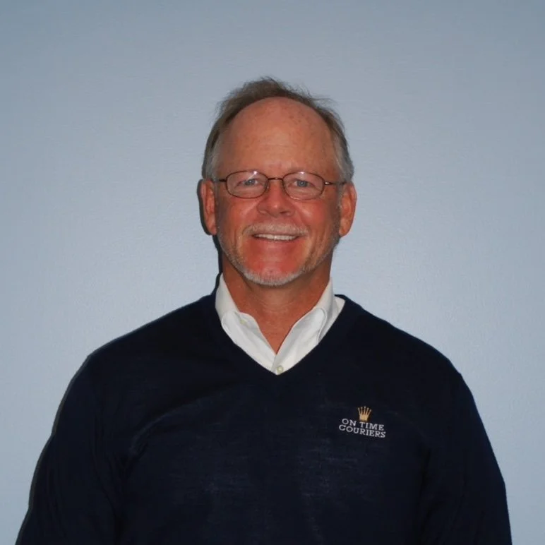 Smiling man with glasses, salt-and-pepper hair, beard, wearing a white shirt and dark sweater with an 'ON TIME COURIERS' logo, standing against a light blue wall.