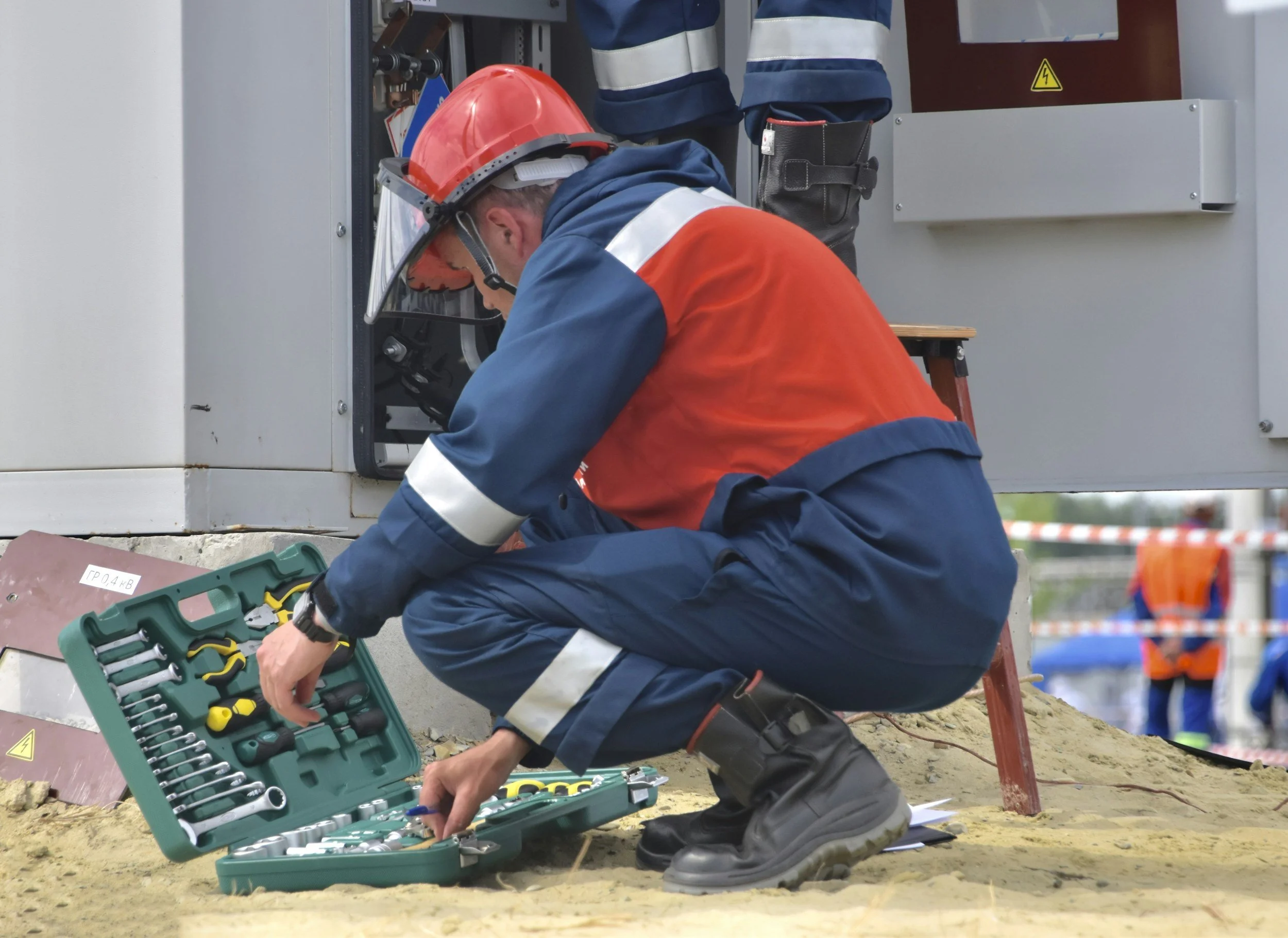 Construction worker kneeling on the ground, selecting tools from a set laid out nearby, wearing a hardhat and safety gear.