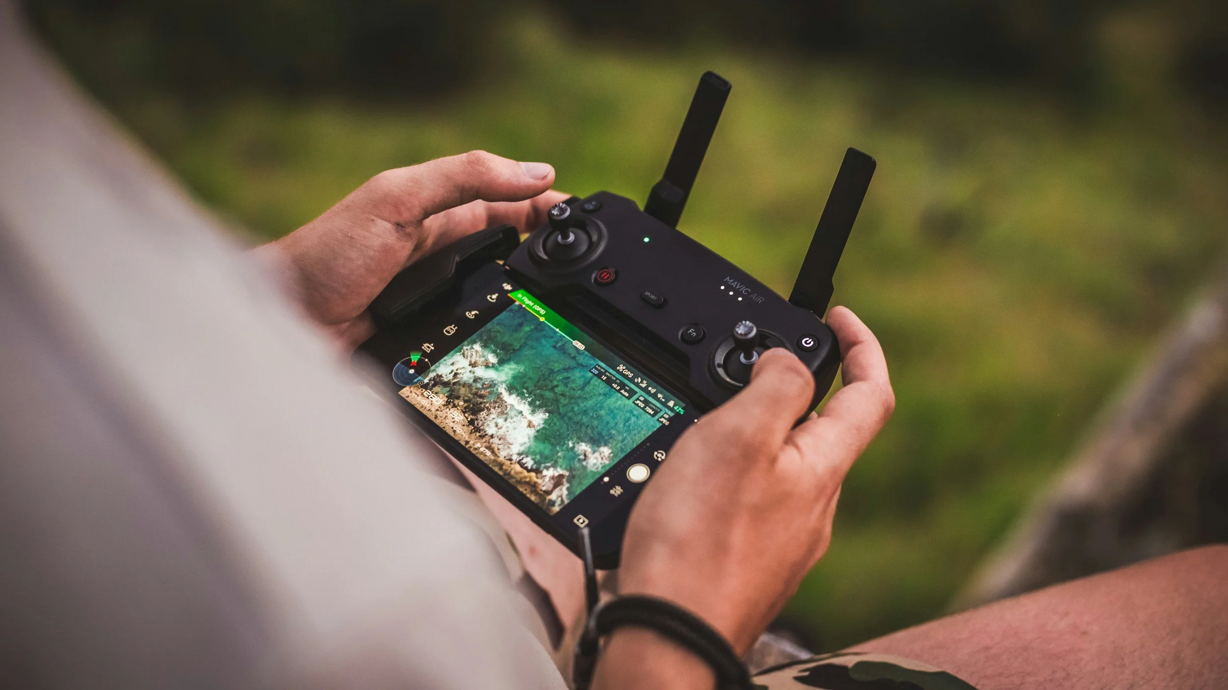 Person holding a drone controller with a built-in screen, actively flying a drone outdoors surrounded by greenery.