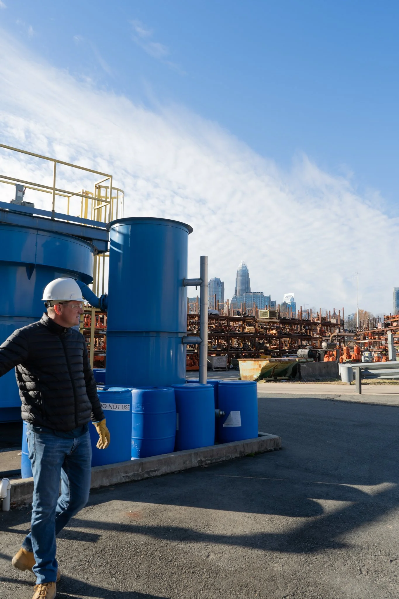 Person wearing a hardhat standing in front of a wastewater treatment plant, with a city skyline visible in the background under a cloudy sky.