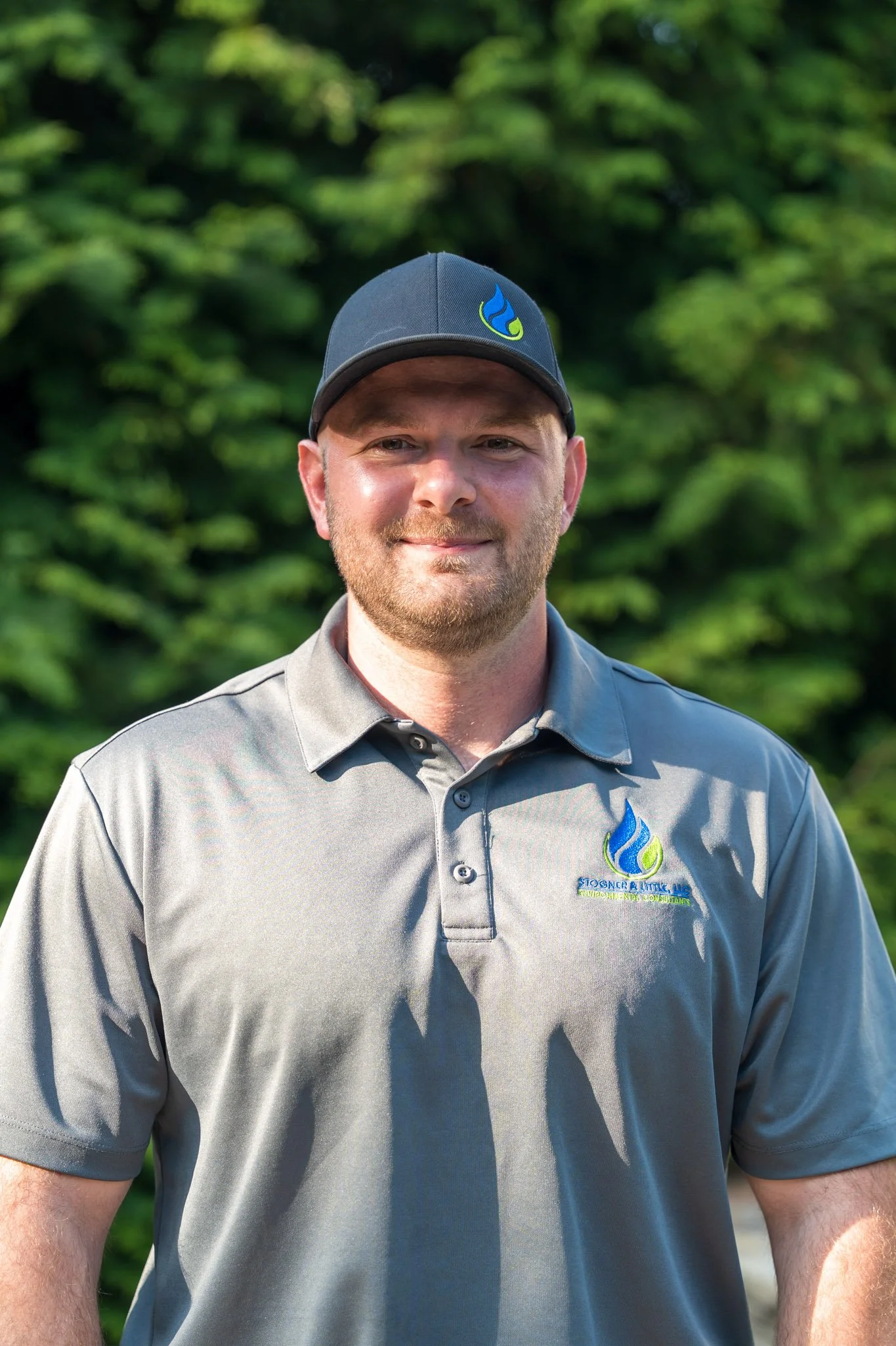 White male wearing a gray polo shirt and a hat, standing and smiling in front of a lush evergreen tree outdoors.