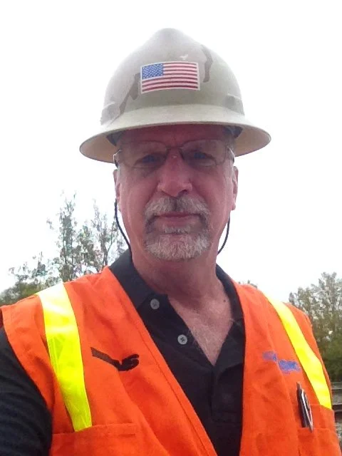 White male wearing a high-visibility orange vest and a camouflage hard hat, standing outdoors in front of a group of trees.