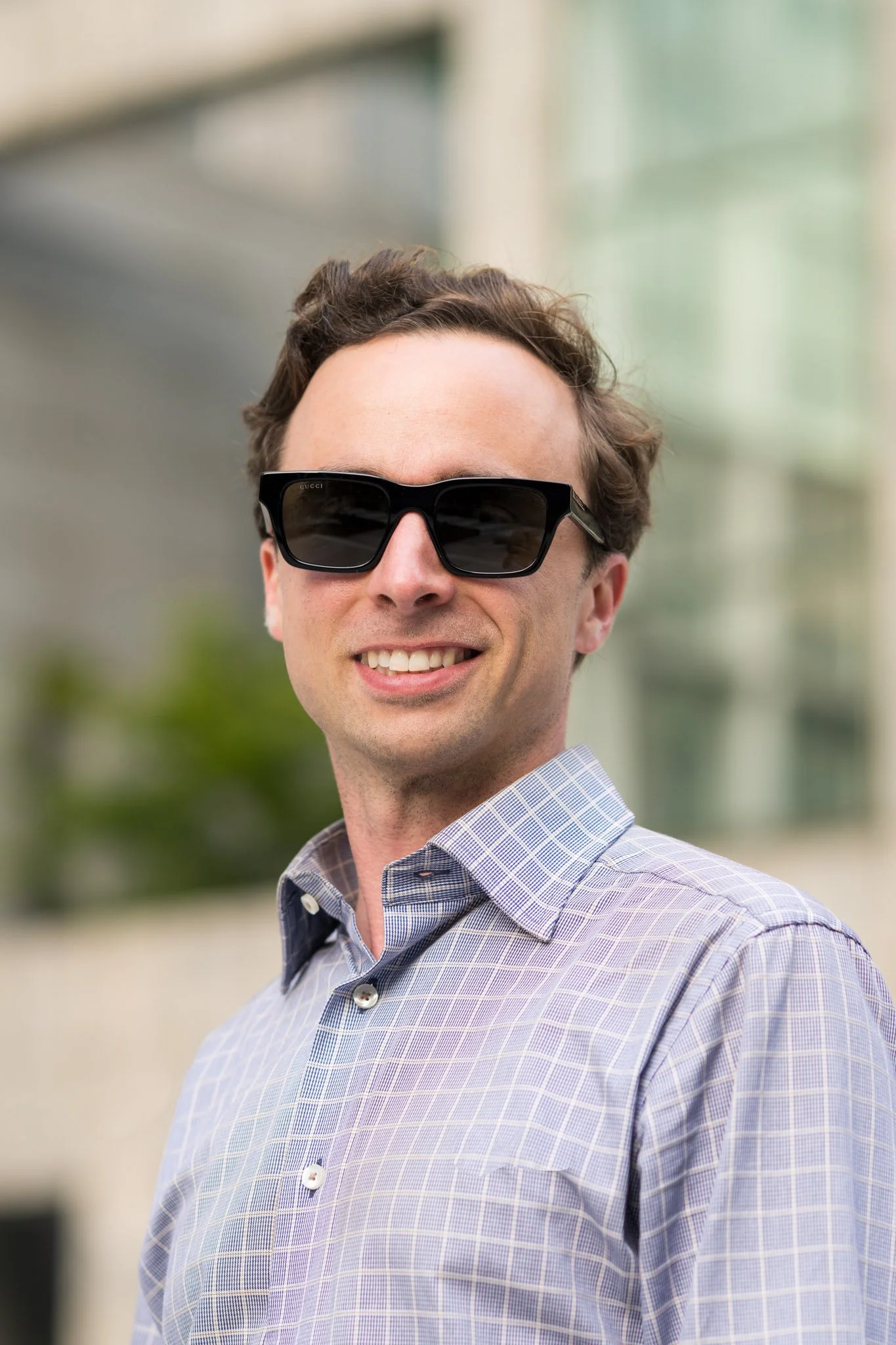 White male wearing a button-up plaid shirt and black sunglasses, smiling while standing in front of a tall skyscraper in an urban setting.