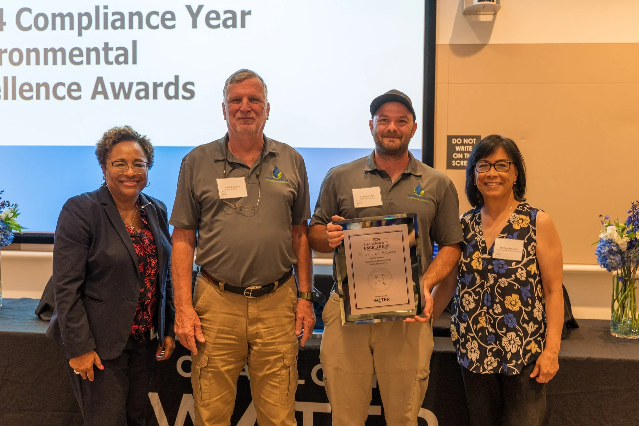 Four people in business casual attire standing in front of a projector screen at an awards banquet, smiling and holding an award for environmental excellence.
