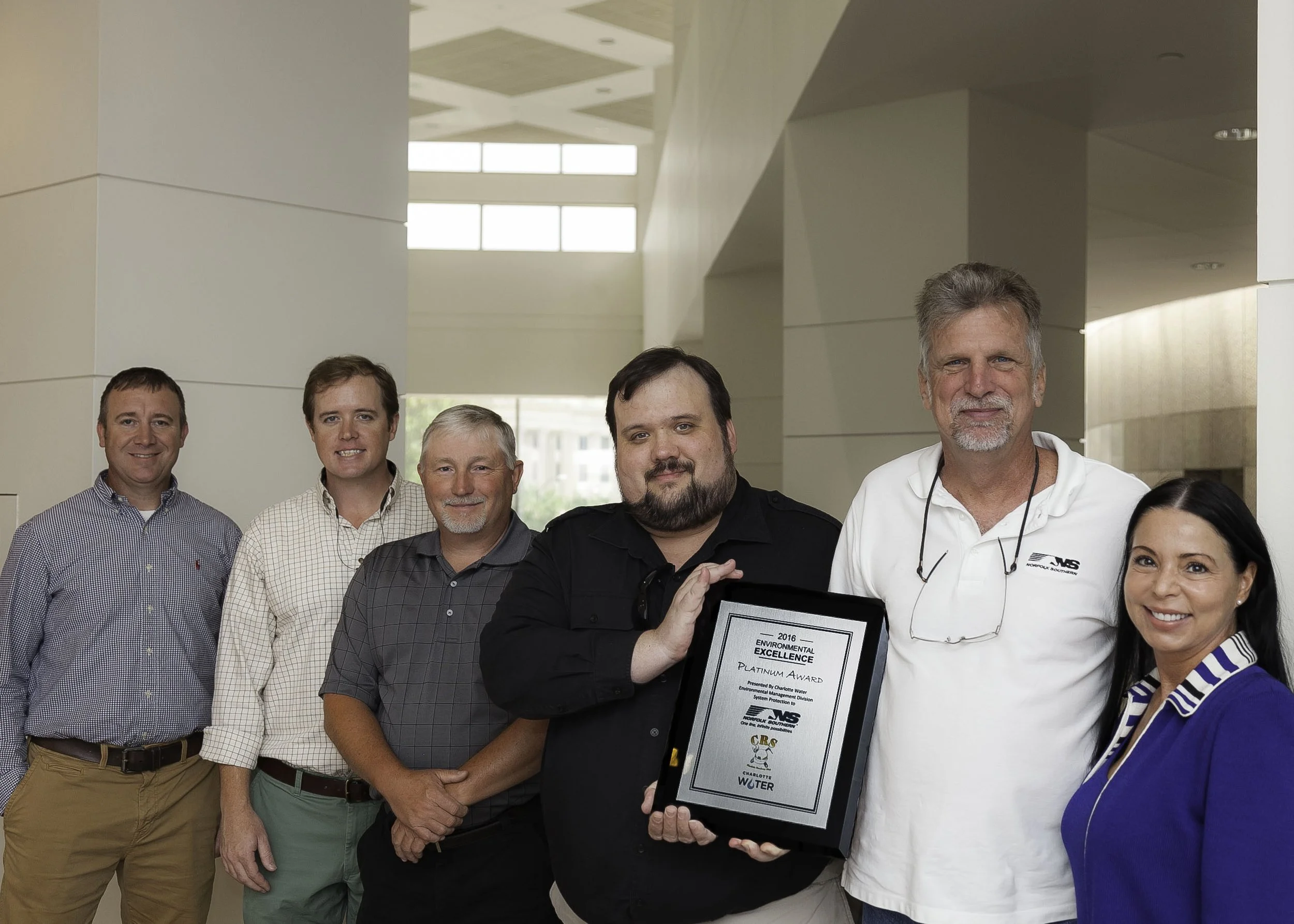 Six people in business casual attire standing together in the lobby of an office building, smiling and holding an award for environmental excellence at an awards banquet.