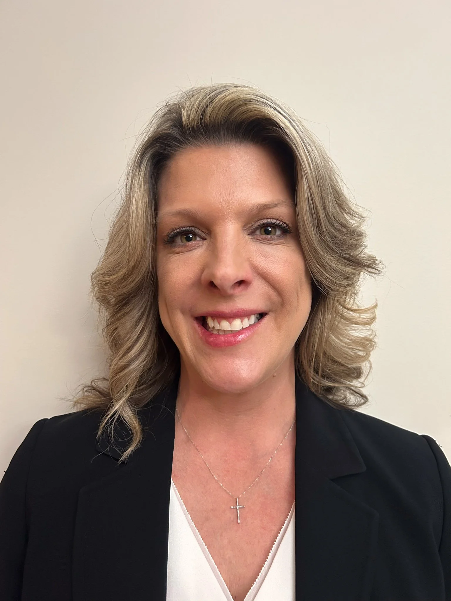 White female wearing a black business suit, standing and smiling in front of an eggshell-colored office wall in a professional indoor setting.