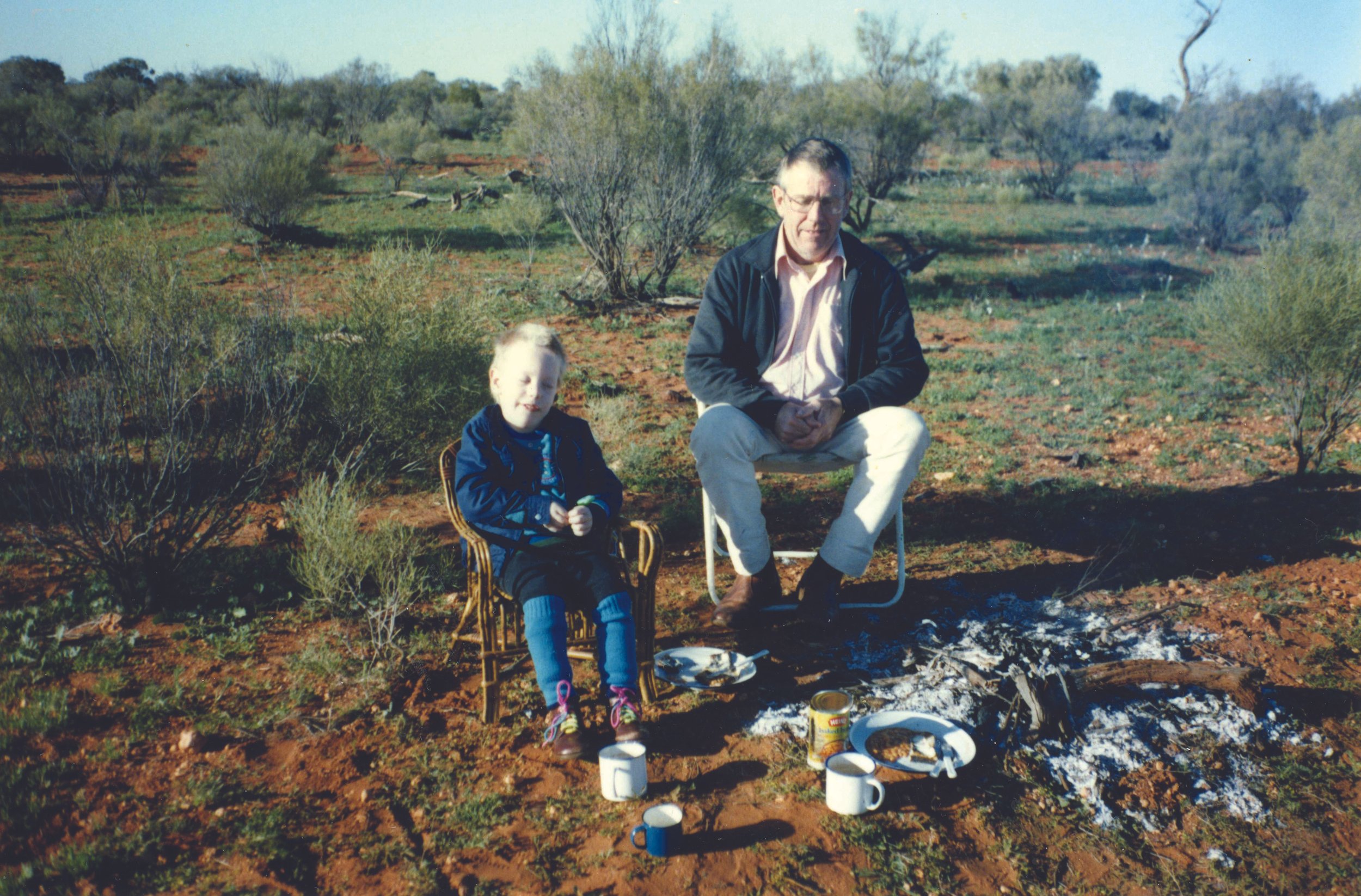 A man and a young girl sitting outdoors by a campfire with plates, mugs, and food, in a dry, bushy landscape with sparse vegetation.