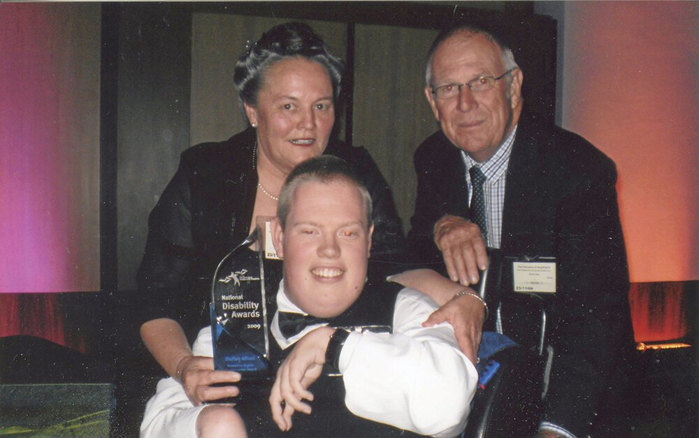 A young man in a wheelchair holding a crystal award, flanked by an older woman and man at an awards ceremony.