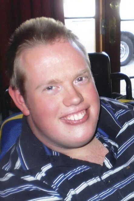 A smiling young man with short light-colored hair, sitting indoors near a window, wearing a blue and white striped shirt.