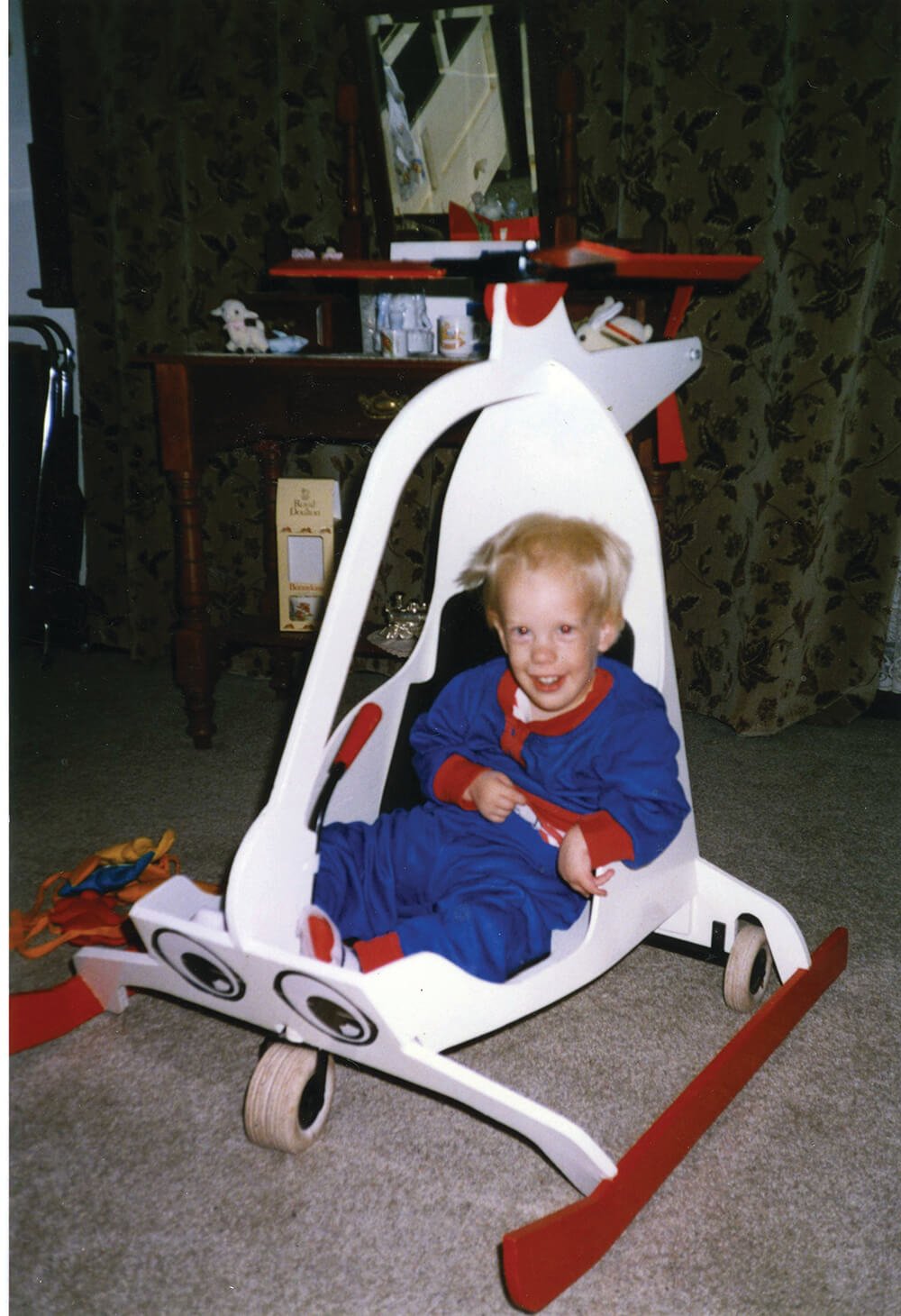 A young child in a blue and red pajamas sitting inside an upside-down toy helicopter with a white body and red accents on a carpeted floor in a living room.
