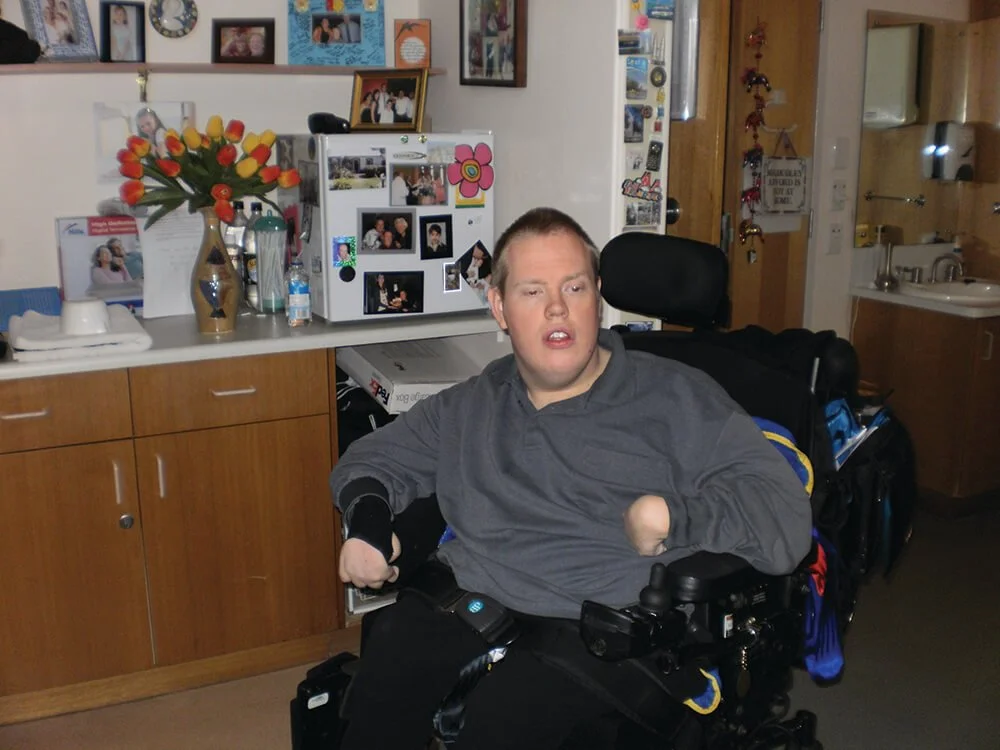 A man with a disability in a motorized wheelchair in a room decorated with photos, a refrigerator with magnets, and a kitchen area in the background.
