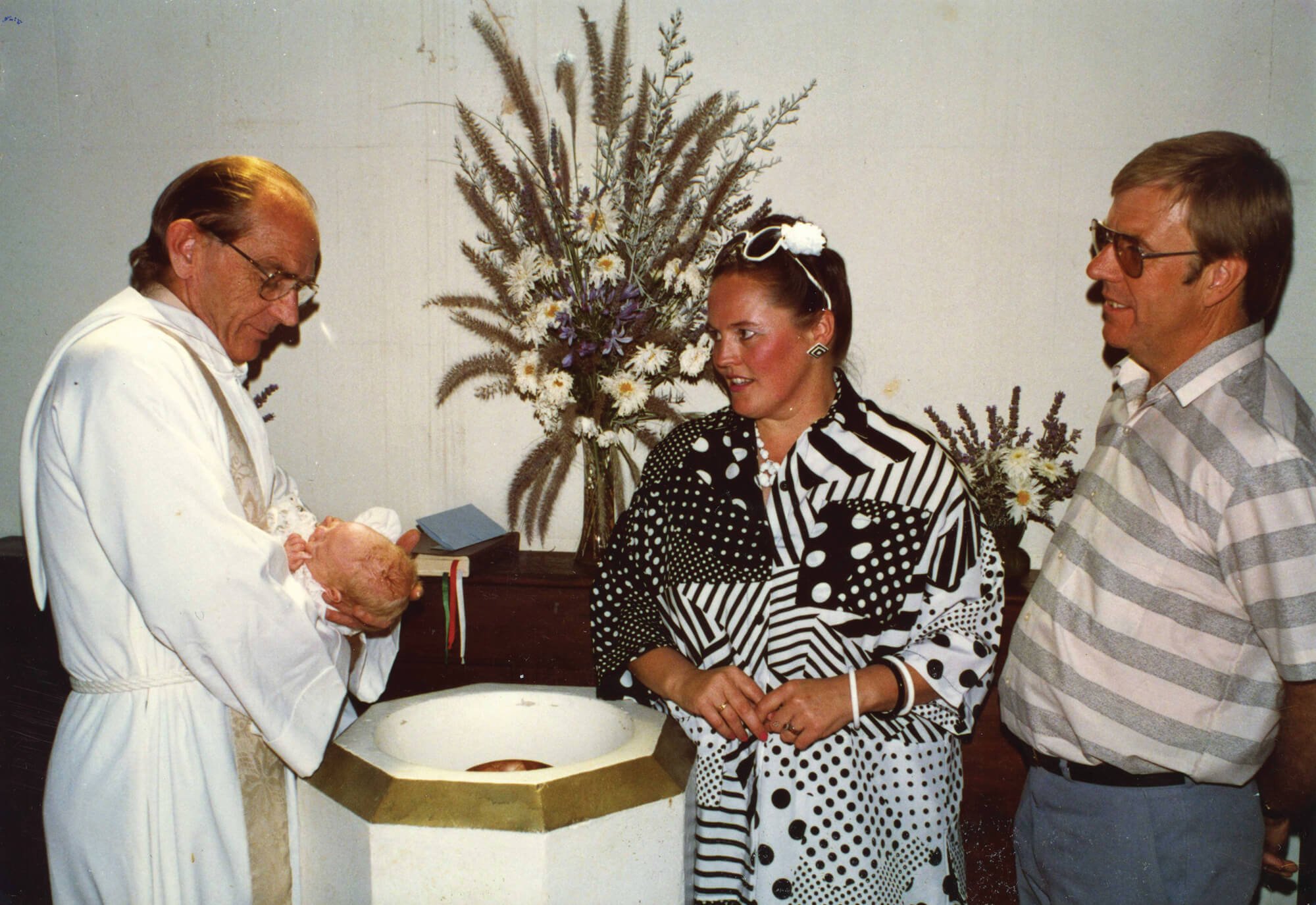 A woman and a man are standing in front of a altar, watching a priest hold a newborn baby in a baptism or christening ceremony. The woman is wearing a black and white patterned dress, and the man is wearing a light striped shirt. The priest is dresse