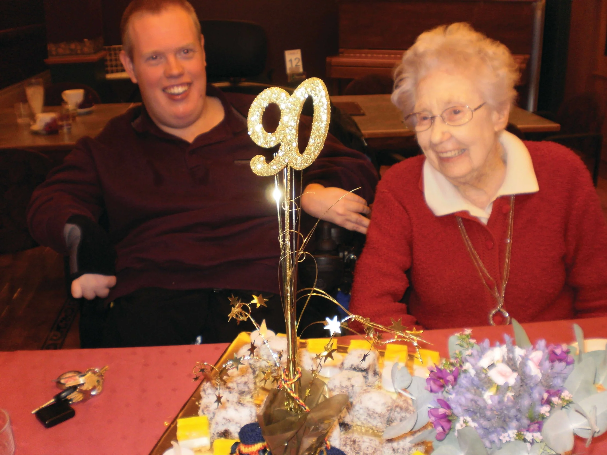 A young man with a disability and an elderly woman celebrating a 90th birthday at a restaurant, with a decorated cake and a gold '90' topper on the table.