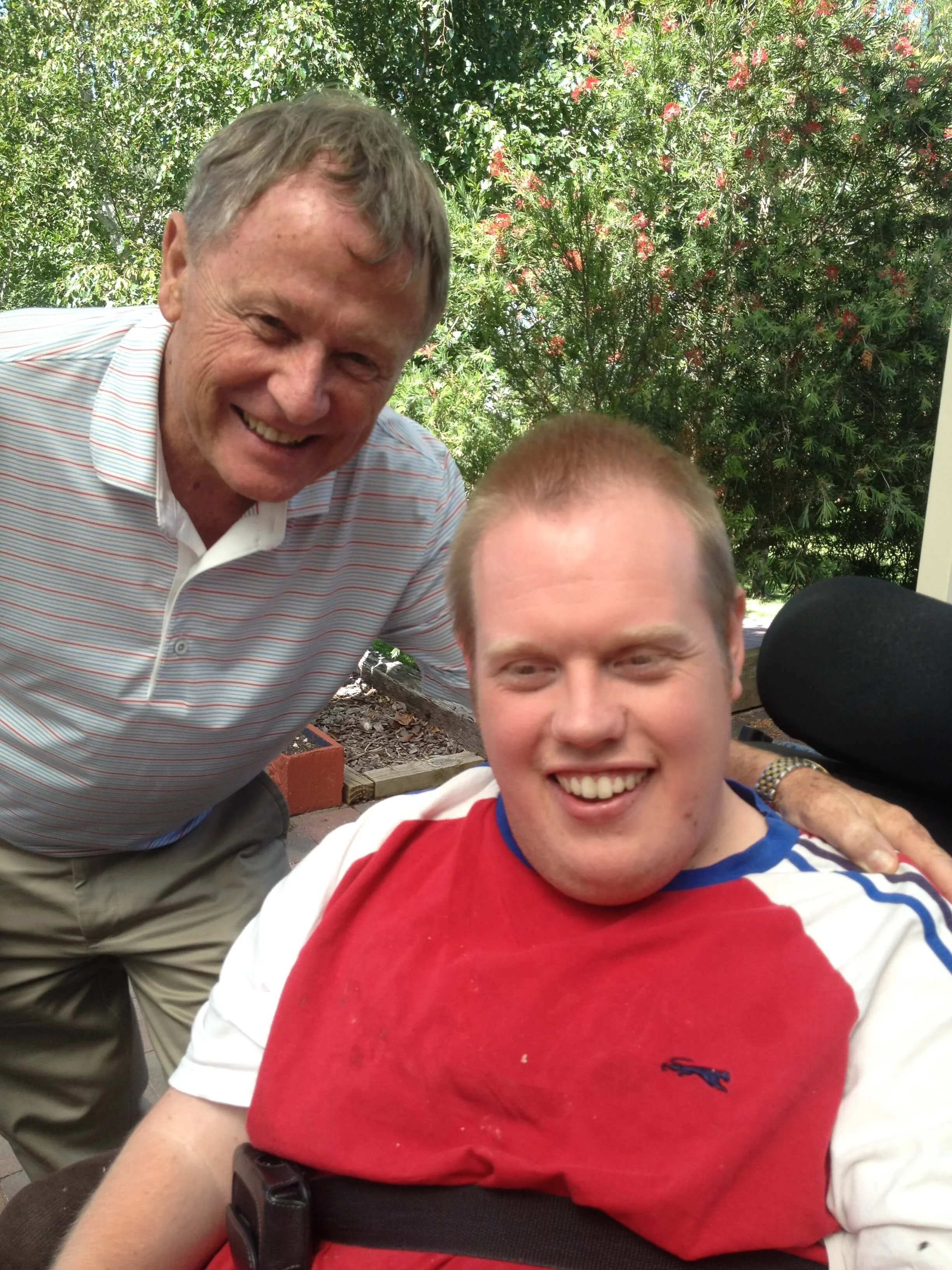 Two men smiling outdoors with greenery in the background, one standing and one sitting in a wheelchair.
