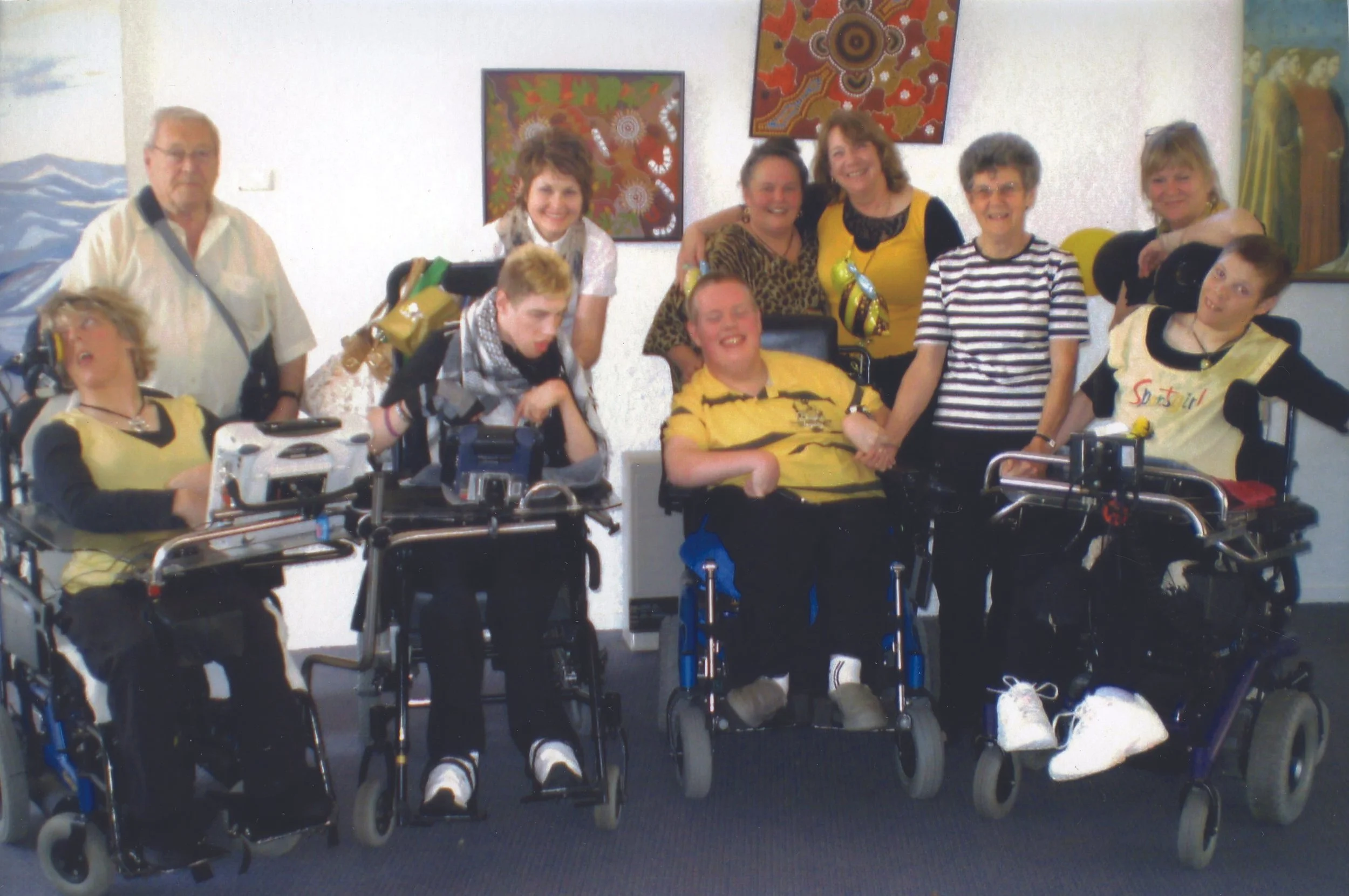 Group of people, some in wheelchairs, smiling and standing together in an art gallery or indoor space with colorful paintings on the wall.