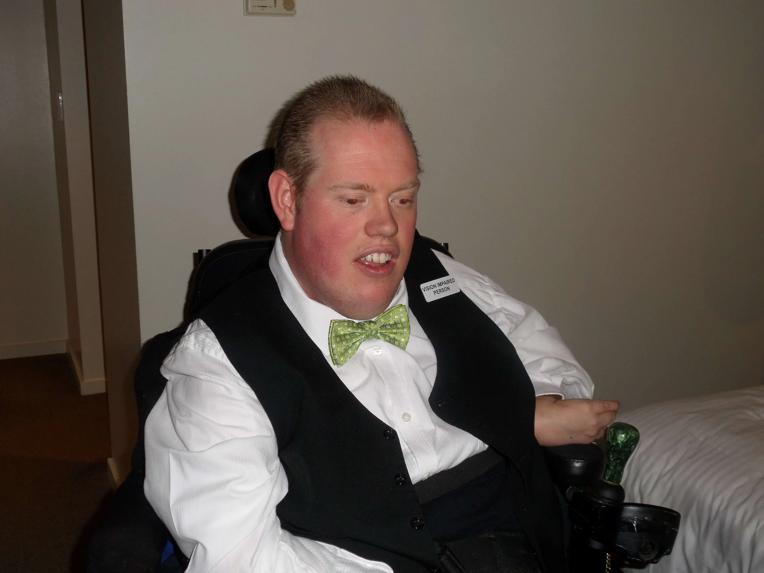 A man in a wheelchair with a spinal cord injury, wearing a white shirt, a black vest, and a green bow tie, sitting indoors against a plain light-colored wall.