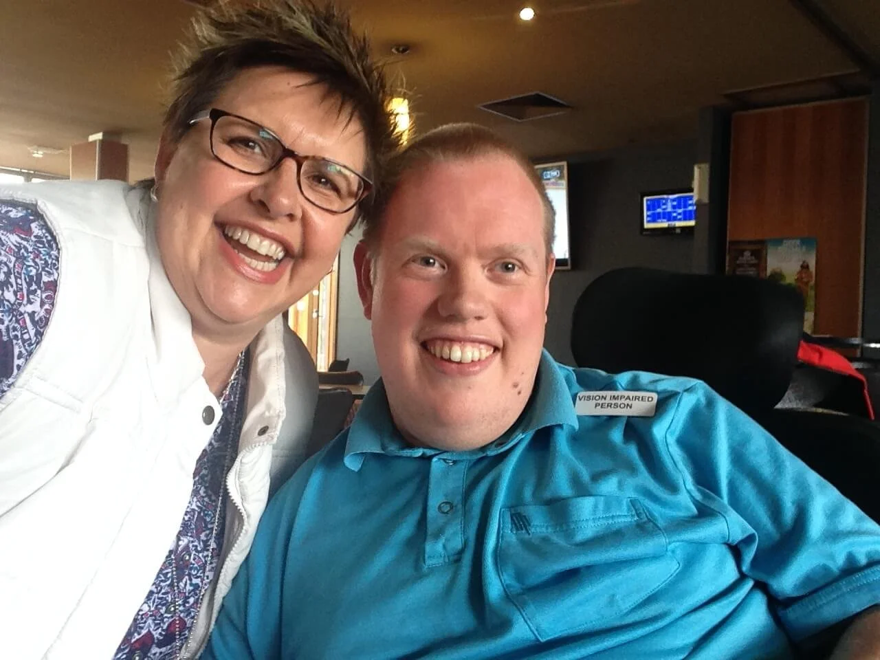 A smiling woman with short dark hair and glasses taking a photo with a smiling man with short light hair, who is in a wheelchair with a label that reads 'Vision Impaired Person', inside a room with TV screens and wooden paneling.