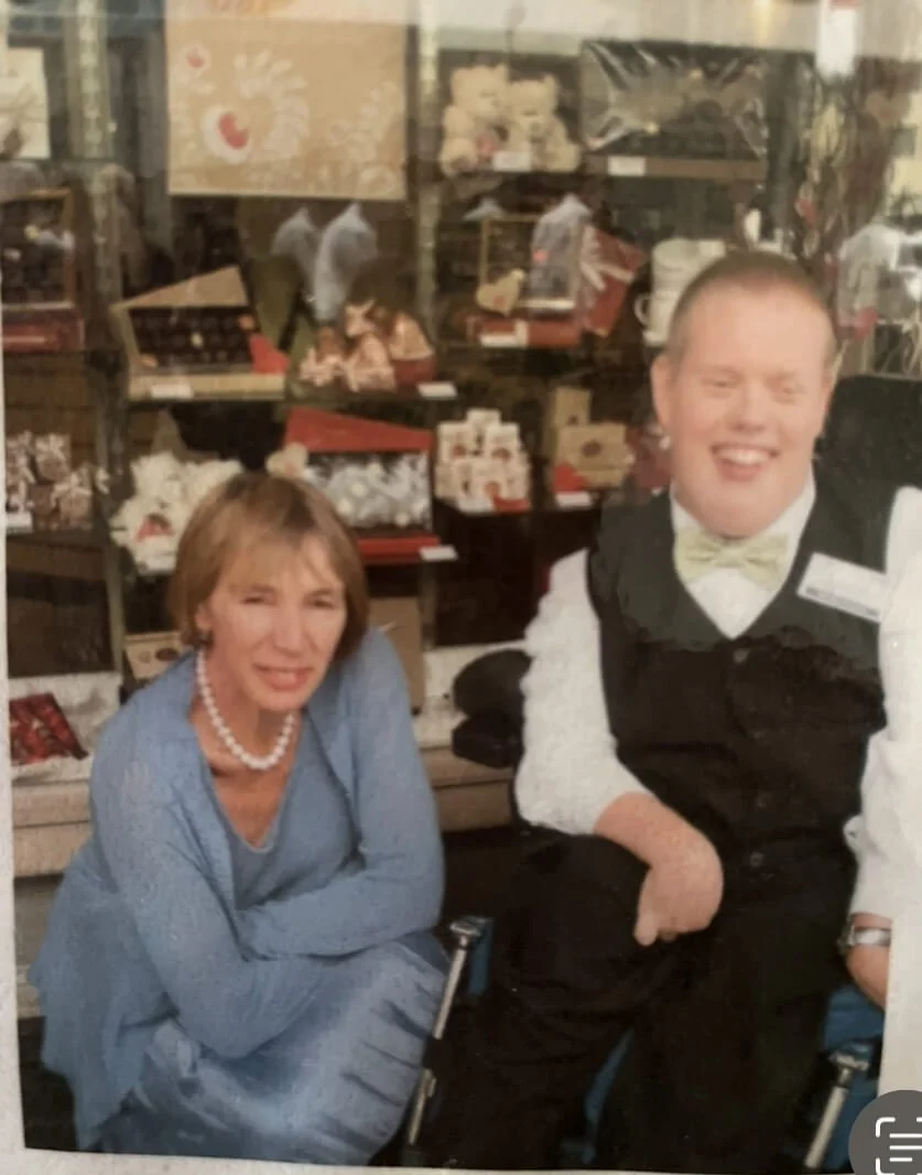 A woman in a blue dress and pearl necklace sitting next to a smiling man in a tuxedo with a bow tie, in a store with shelves of various items behind them.