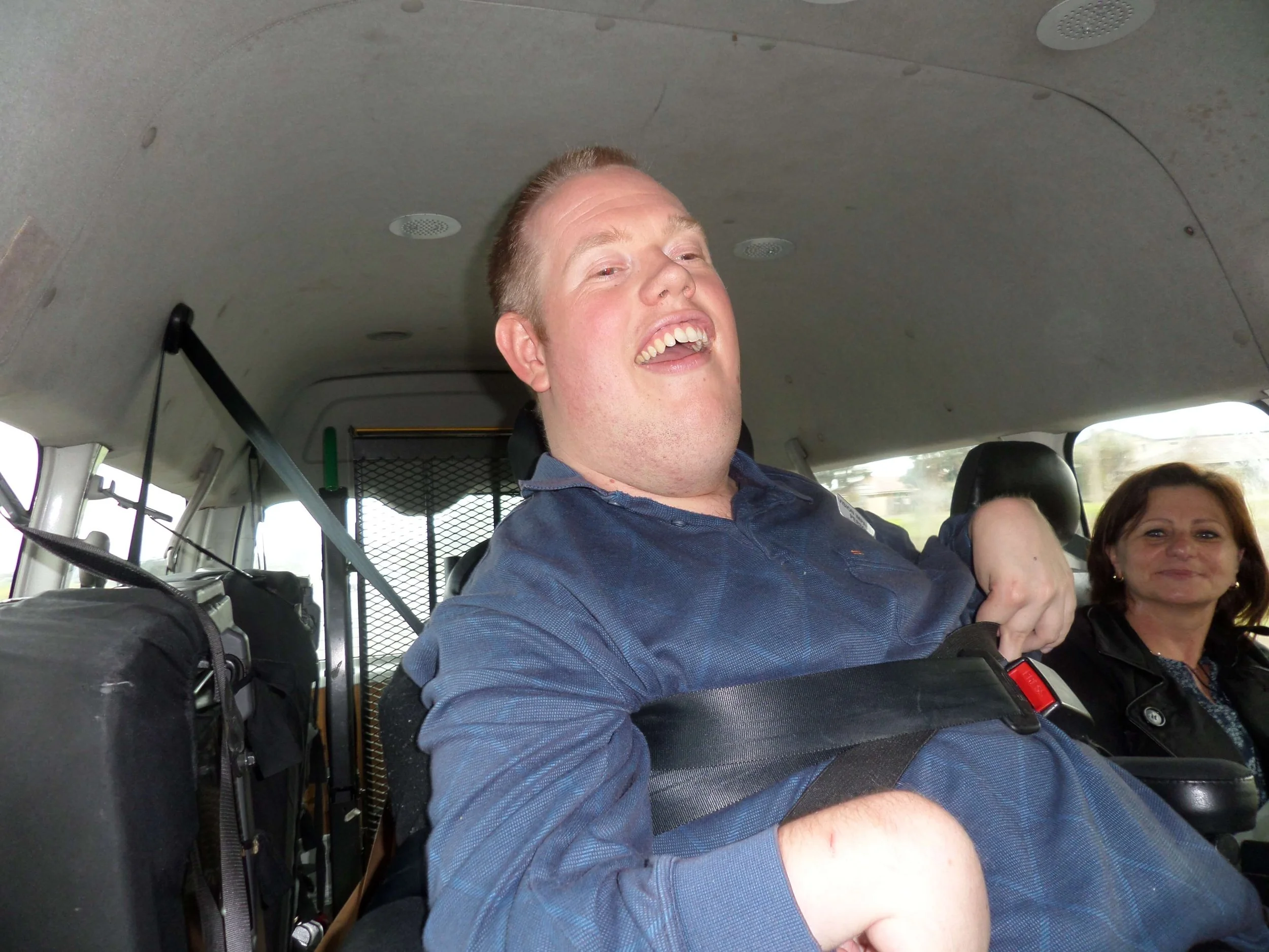 Man smiling and laughing in the backseat of a vehicle, with a woman sitting behind him.