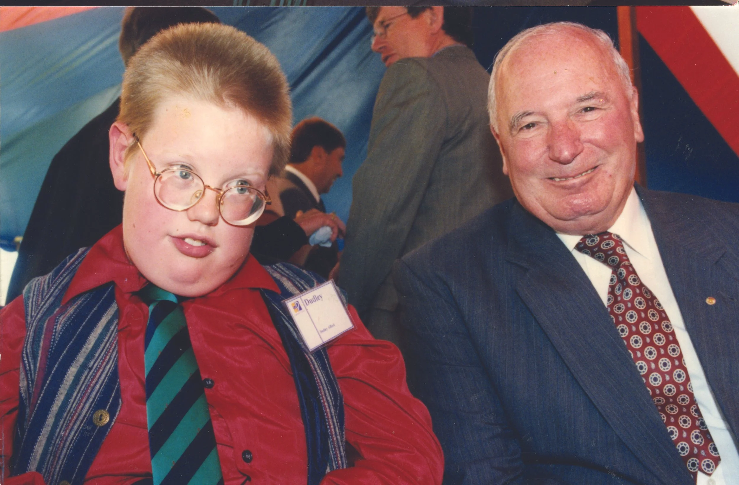 A young man with glasses wearing a red jacket, striped shirt, and tie sits next to an older man in a suit and patterned tie at an event.