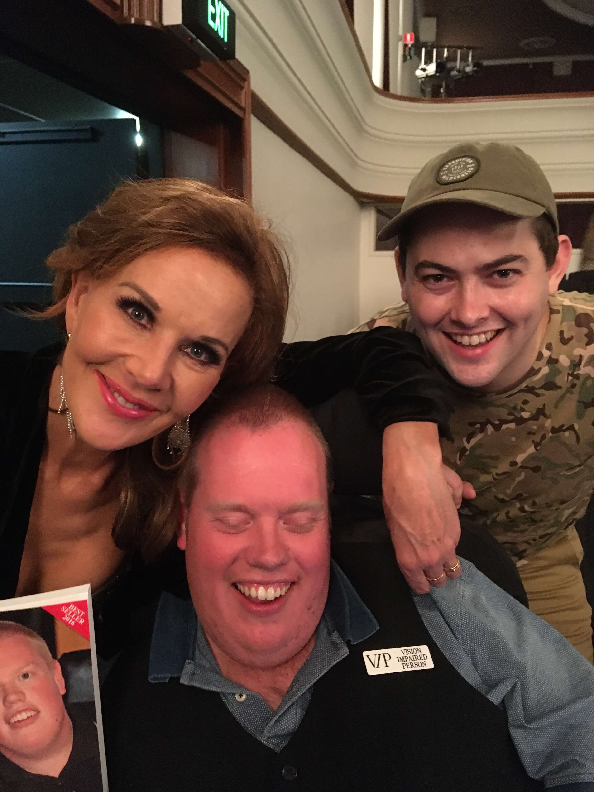 Three people smiling and posing for a photo together indoors, with one person in a wheelchair holding a small photo, and another wearing a camouflage shirt and cap.