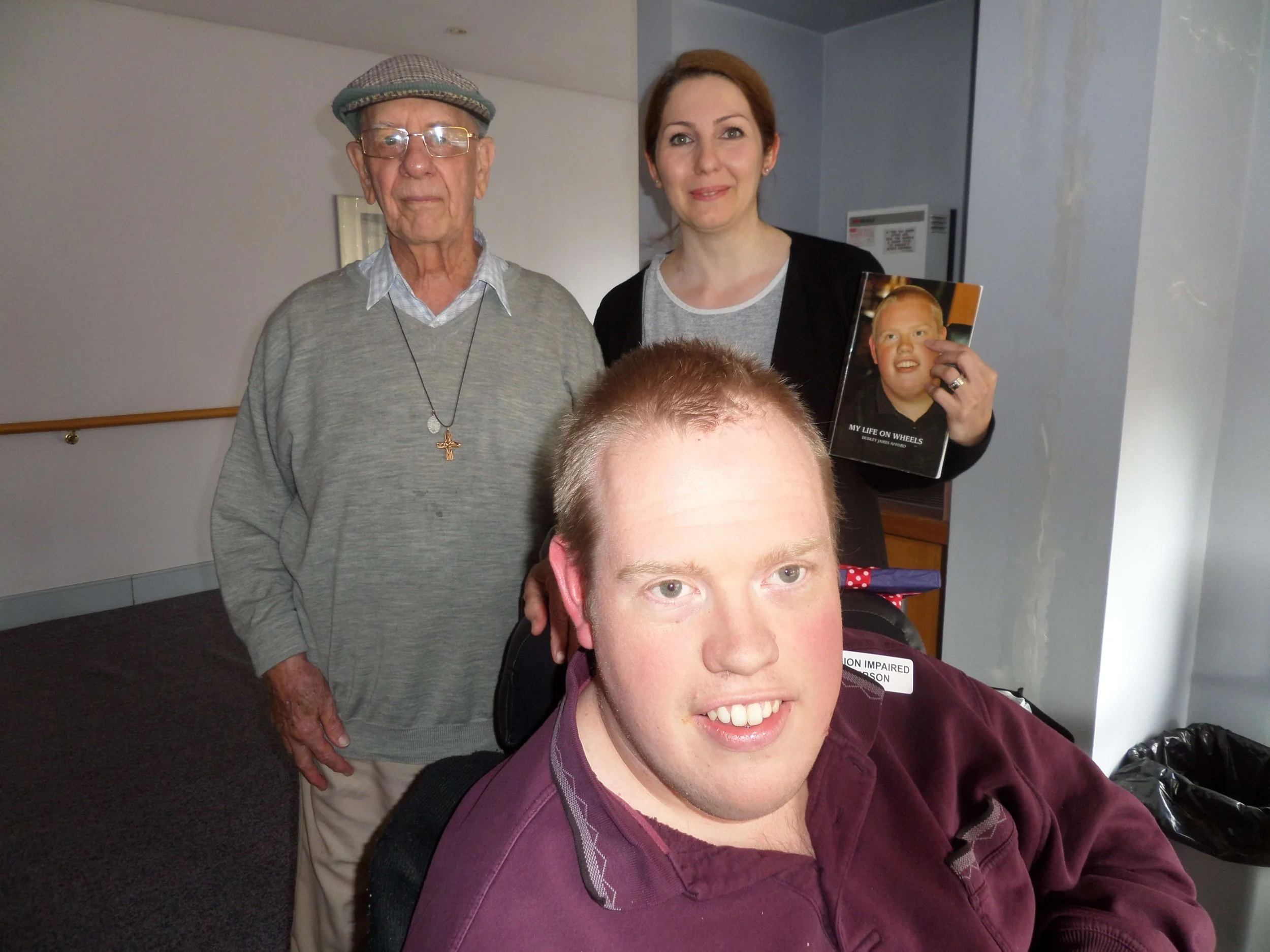 Four people, including a young man in a wheelchair, two women, and an elderly man, pose for a photo indoors. The young man is smiling in the foreground, the elderly man wears glasses and a flat cap, and the women stand behind them with one holding a 