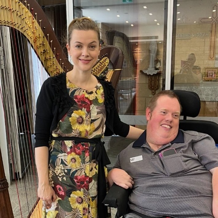 A woman and a man with a disability smiling for a photo inside a building with a large harp in the background.