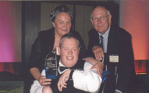 A young man in a wheelchair being honored with a Disability Awareness Award, surrounded by two older adults, a woman and a man, at an award ceremony.