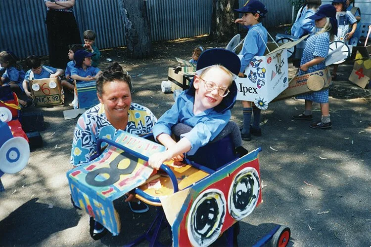 Children and adults participating in a school event with decorated carts and costumes outside, some children sitting in and riding on handmade carts.