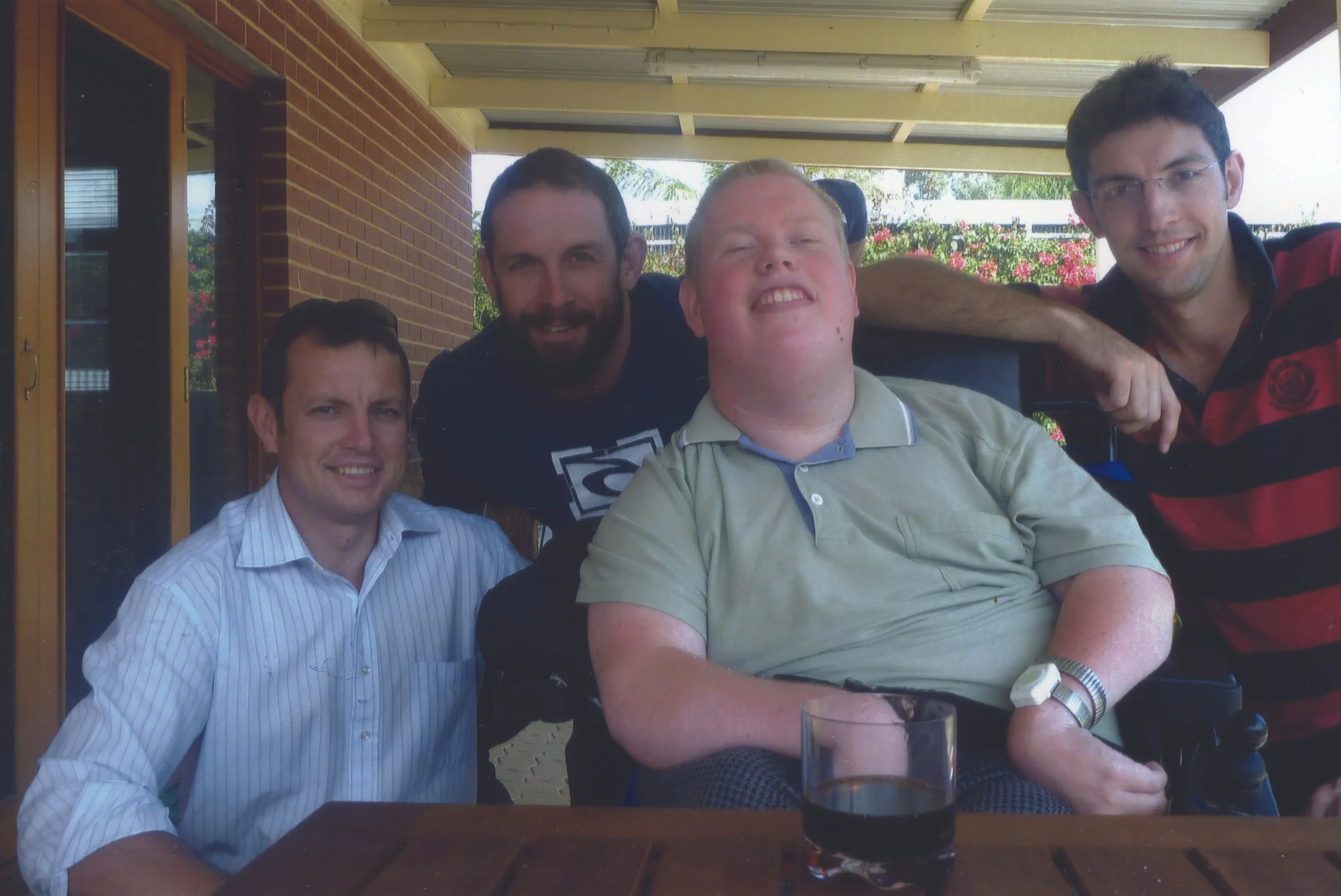 Four men smiling and posing for a photo on a covered patio with a brick wall behind them and trees in the background. One man in a wheelchair is in the center, with three others standing or sitting around him.