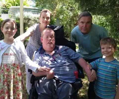 Five people, including two children and three adults, smiling outdoors with trees and a fence in the background.