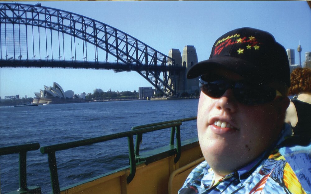 Person wearing a black cap with red and yellow embroidery, sunglasses, and a colorful Hawaiian shirt taking a selfie with the Sydney Opera House and Harbour Bridge in the background, on a ferry in Sydney, Australia.