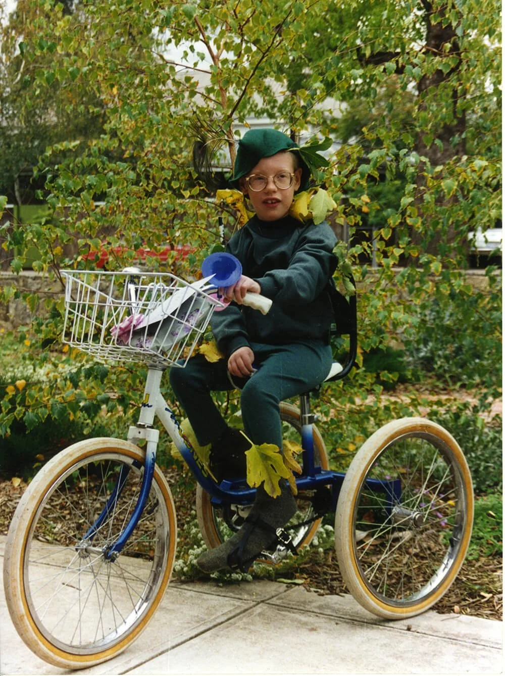 Young girl with glasses riding a bicycle with a basket in front, wearing a dark jacket and green hat, surrounded by leaves and trees in an outdoor setting.