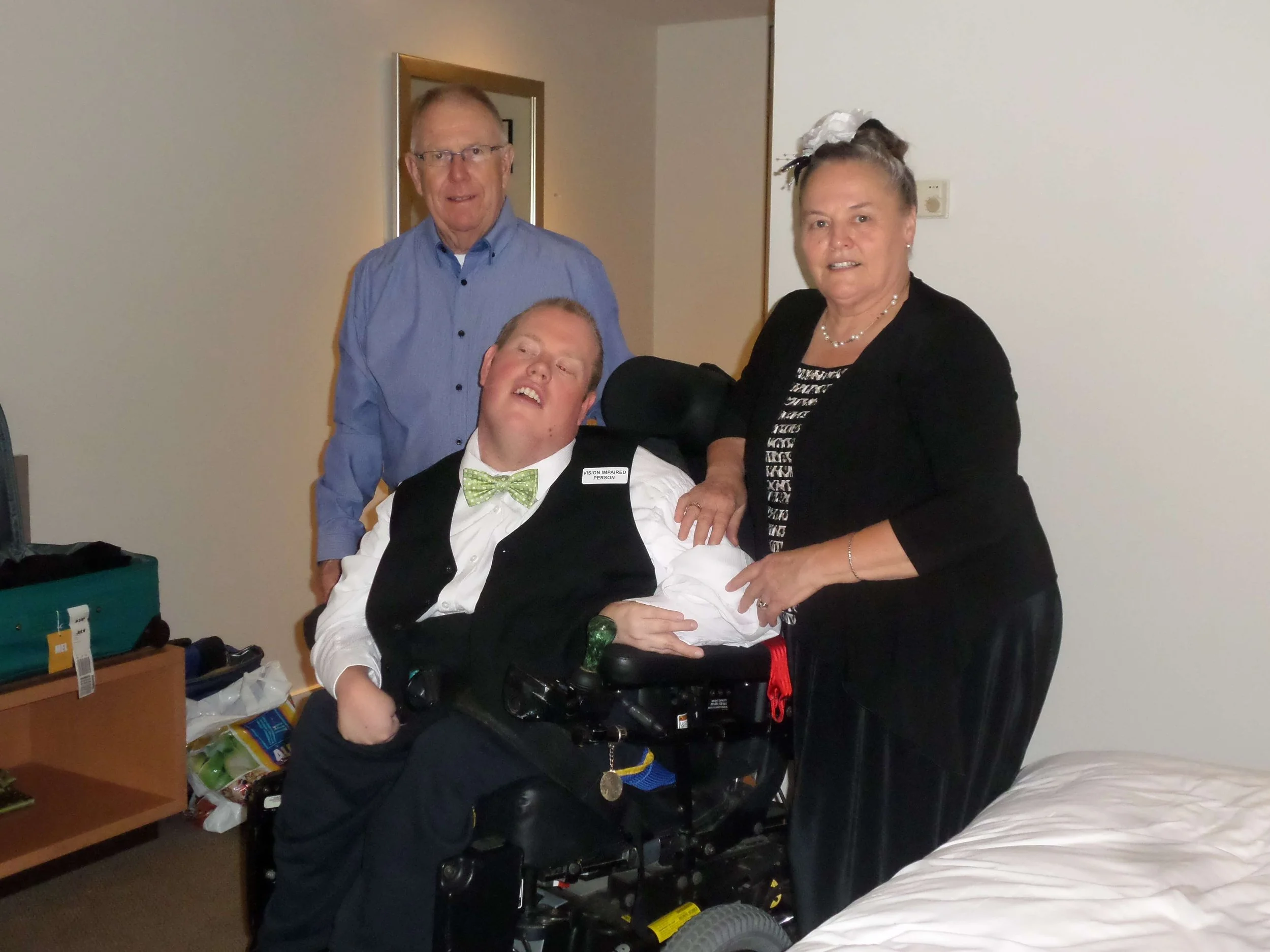 Four people pose in a room, one in a wheelchair, with a bed in the foreground. The person in the wheelchair wears a white shirt, black vest, and green bowtie. An older man in a blue shirt and glasses, an older woman in a black outfit with striped top