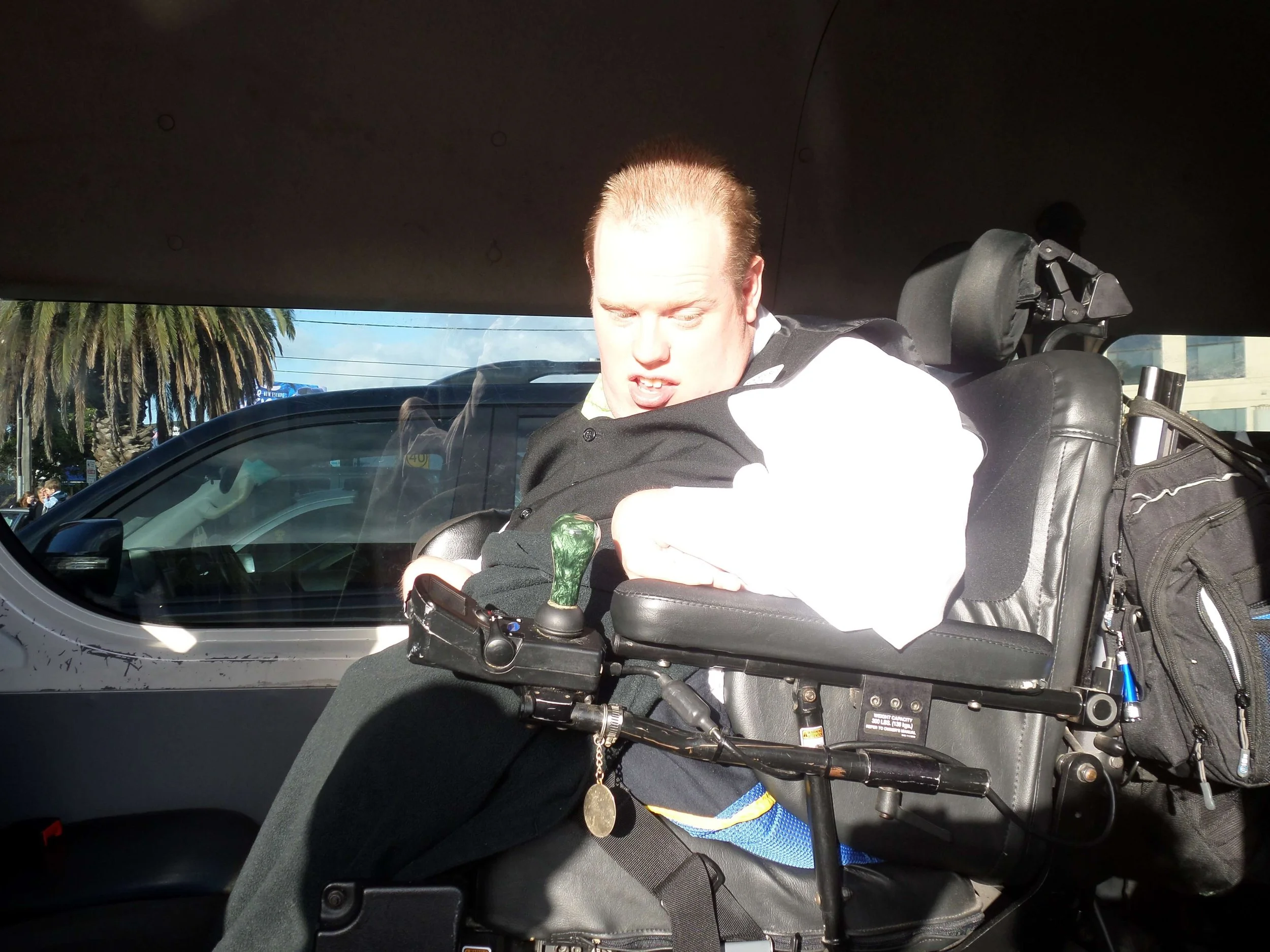 Person in a motorized wheelchair looking down at a mobile device inside a vehicle with palm trees outside through the window.