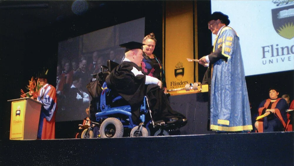A graduation ceremony at Flinders University featuring a person in a motorized wheelchair receiving a diploma from a university official in academic robes, with other graduates and faculty in the background.
