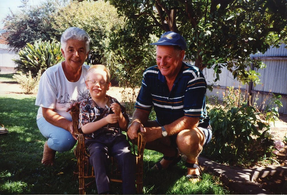 A smiling elderly woman, a young boy, and an elderly man posing in a backyard garden with trees and bushes, the boy sitting on a small wooden chair.