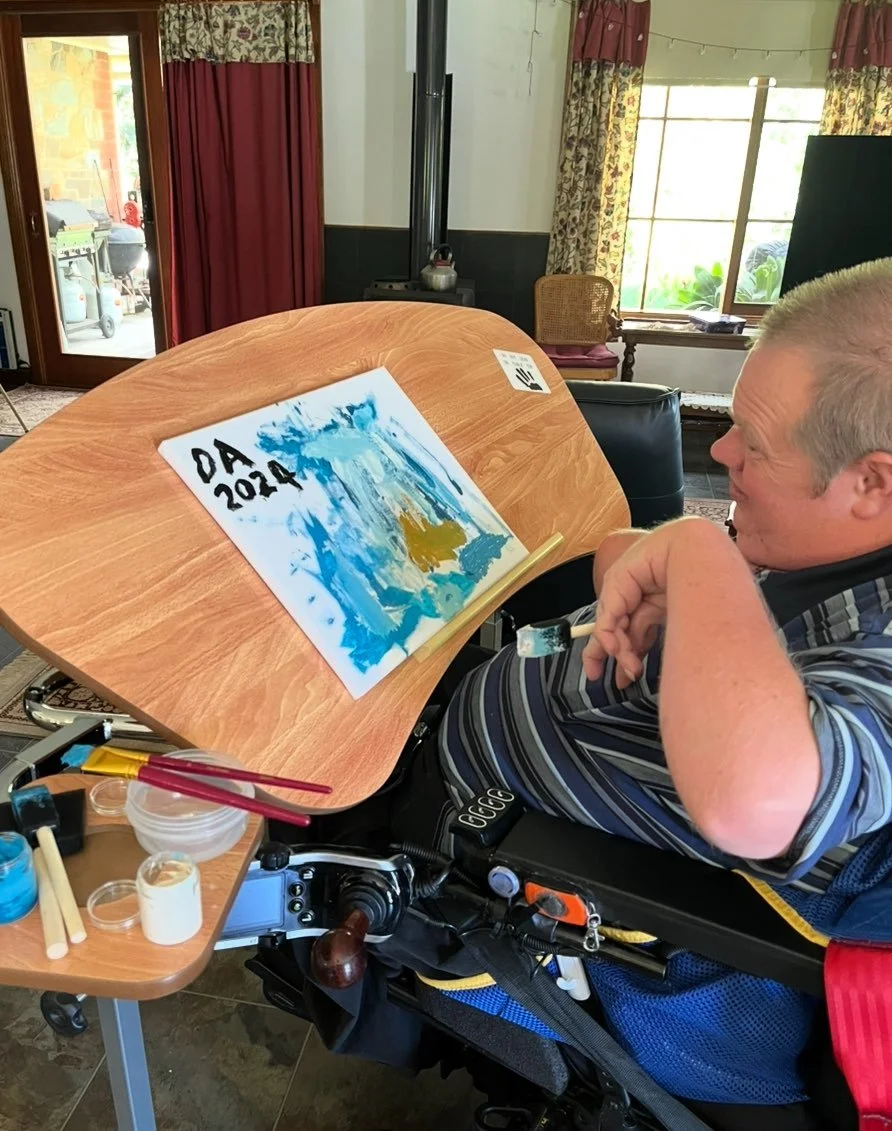 A man in a wheelchair painting on a canvas at a wooden easel inside a room with floral curtains and a large window. The painting has blue, gold, and white brushstrokes, and the words 'DA 2024' written in the top left corner.