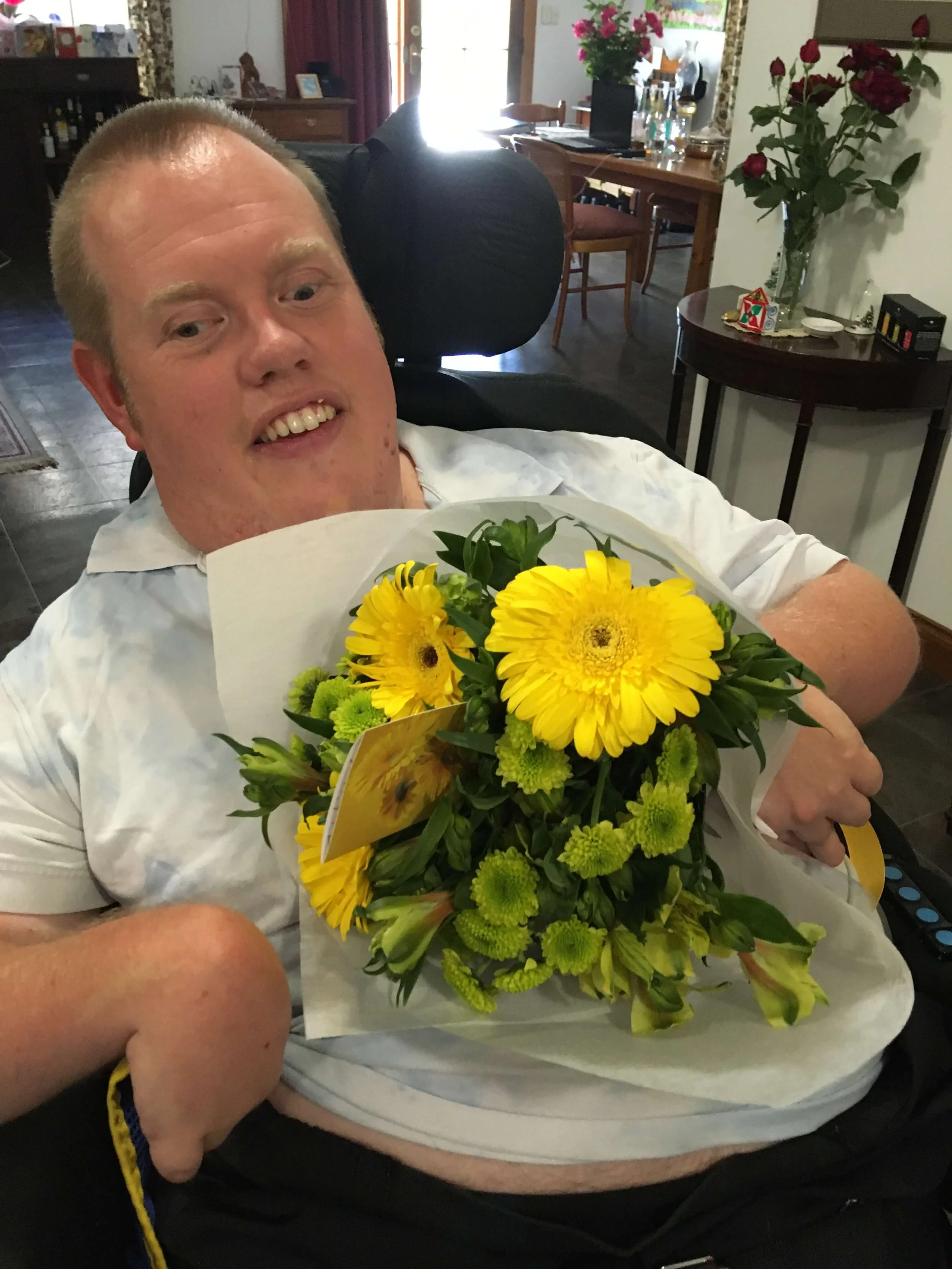 A man in a white shirt sitting in a wheelchair, holding a yellow flower bouquet, smiling.