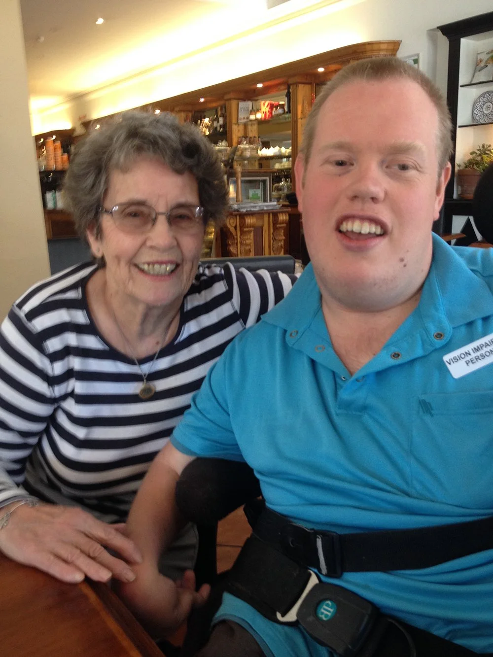 A smiling elderly woman with gray hair and glasses and a younger man with short light hair are taking a selfie together inside a restaurant or cafe. The woman is wearing a black and white striped top and a necklace, and the man is wearing a bright blue shirt with a disability badge and a waist pack. They appear happy and are sitting at a table with a wooden counter and kitchen area in the background.