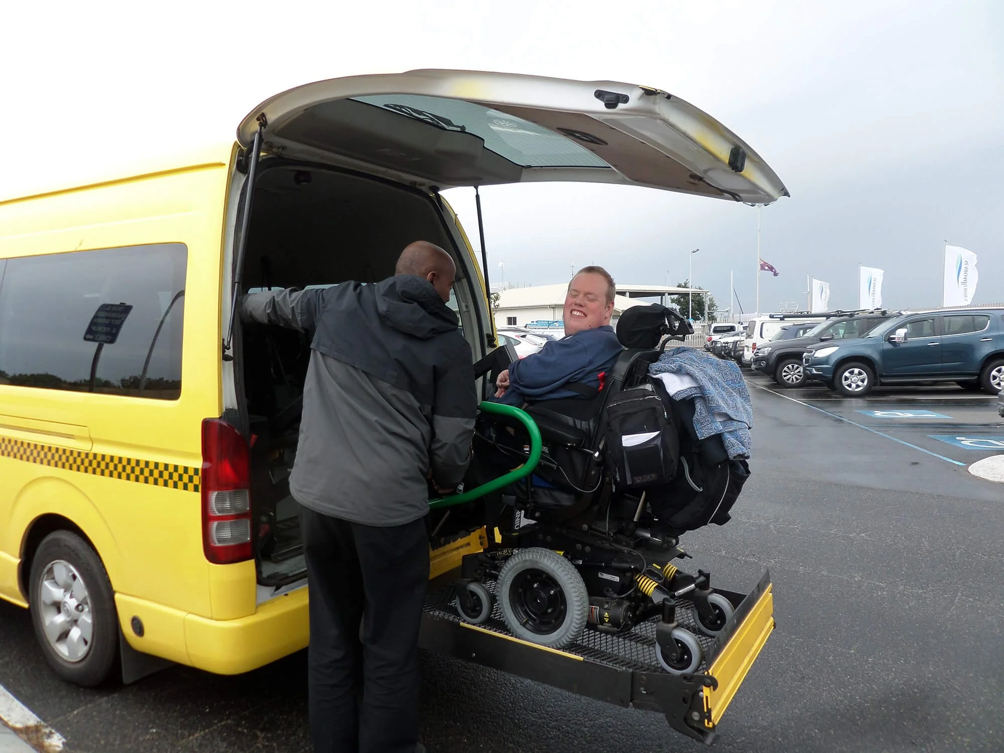A person in a wheelchair being loaded into a yellow transportation van by a staff member in a parking lot during a cloudy day.