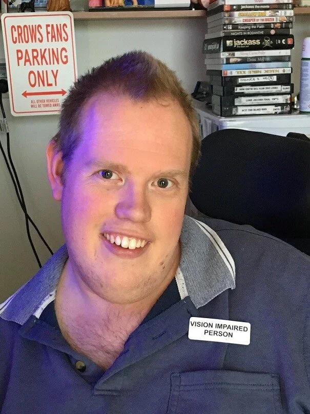 A smiling man in a polo shirt with a badge that says 'Vision Impaired Person,' sitting in an office or living room with a stack of DVDs and a sign that reads 'Crows Fans Parking Only' in the background.