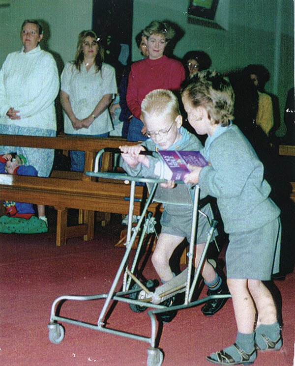 Two young boys using a walker together, looking at a magazine, in a room with several people standing and sitting in the background.