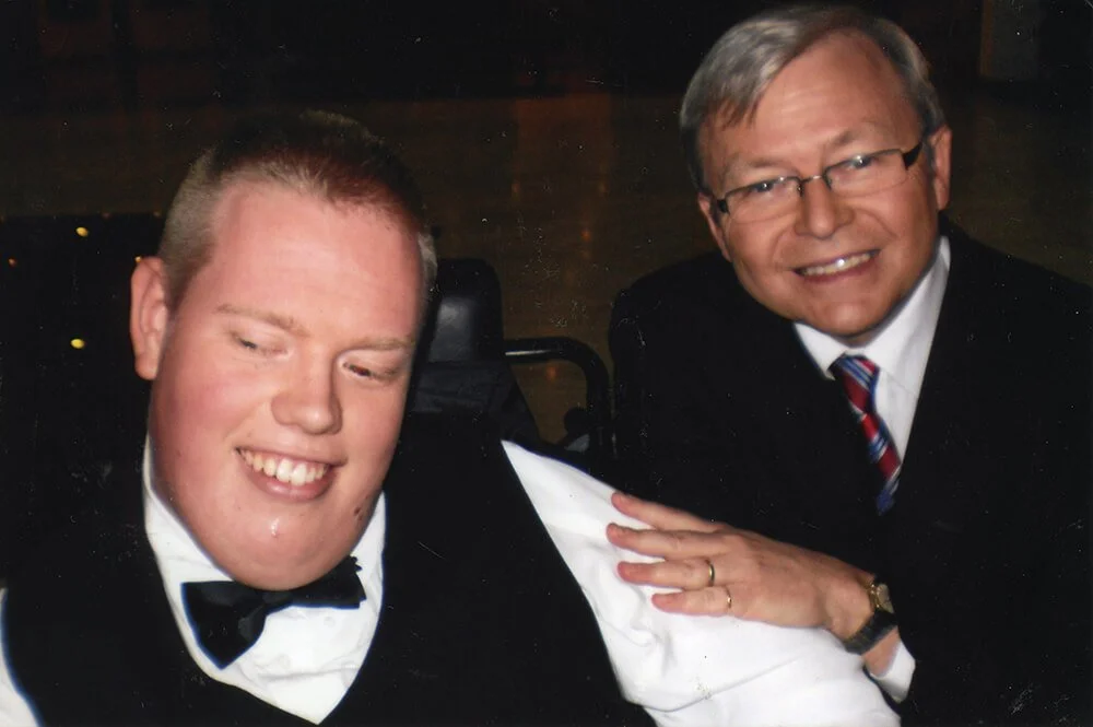 Two men in formal attire, one with glasses, smiling, indoors with dark background.